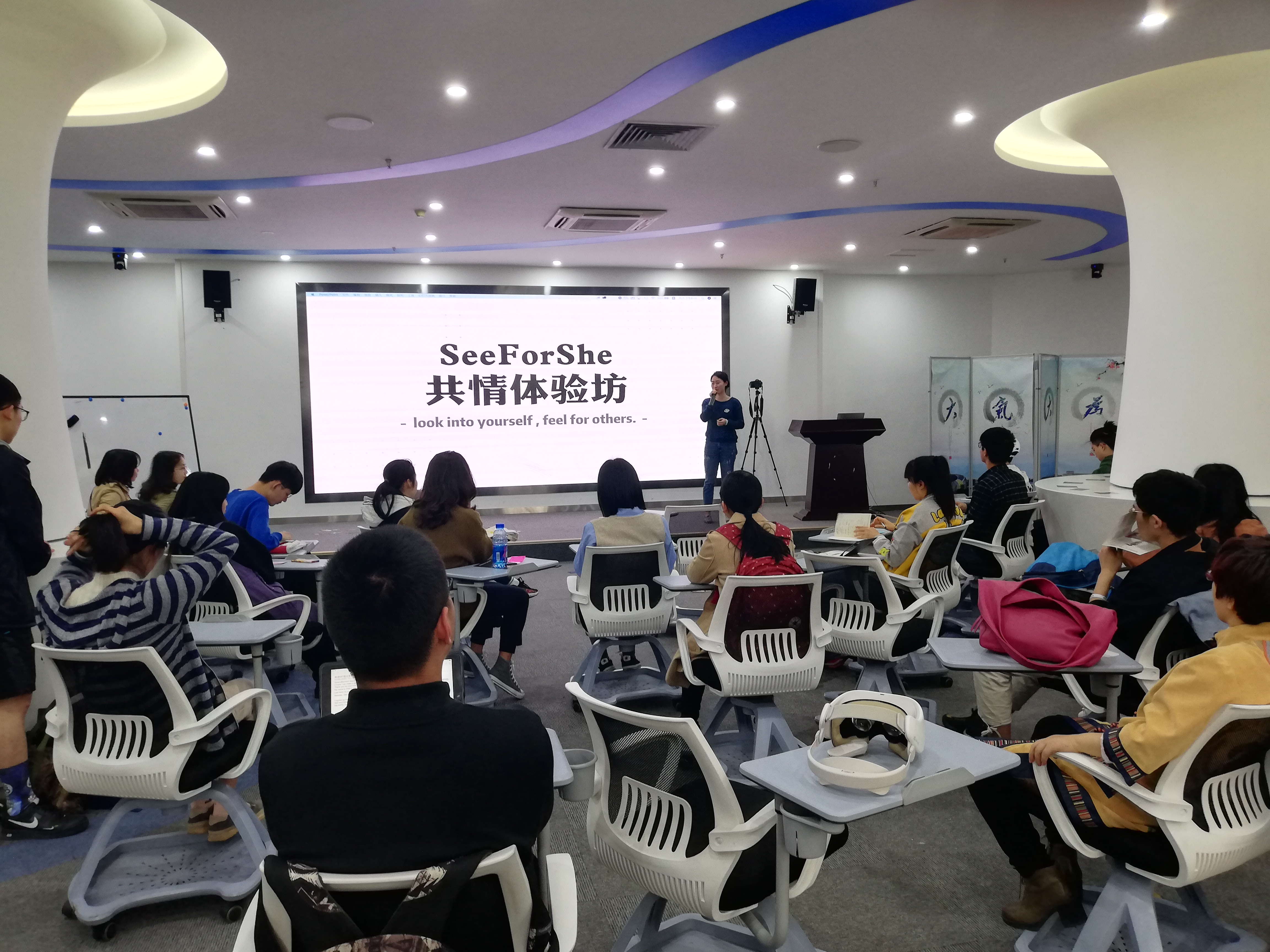 Workshop scene for SeeForShe, showing participants seated in a classroom while a facilitator introduces the empathy experience in front of a large screen.